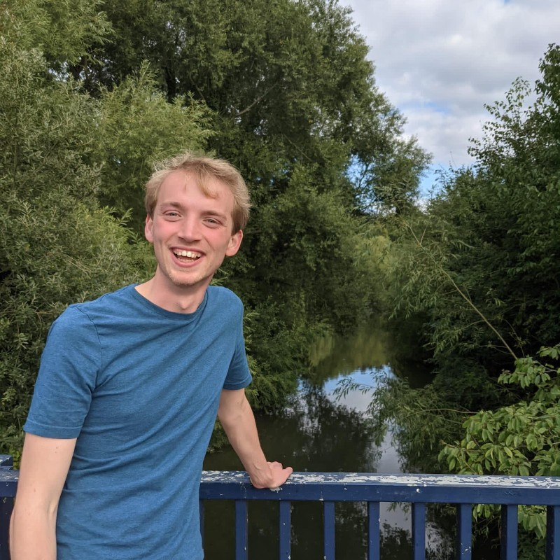 Smiling person in a blue shirt leaning on a bridge railing by a river under cloudy skies