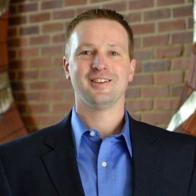 Man in a dark blazer and blue shirt smiling in front of a brick wall