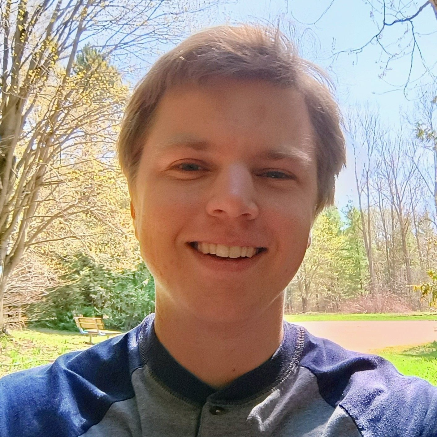 Smiling person outdoors in a sunny park, with trees and blue sky behind them