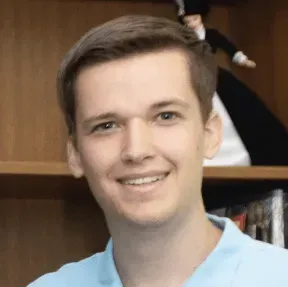 A man in a blue shirt is smiling in front of a bookshelf.