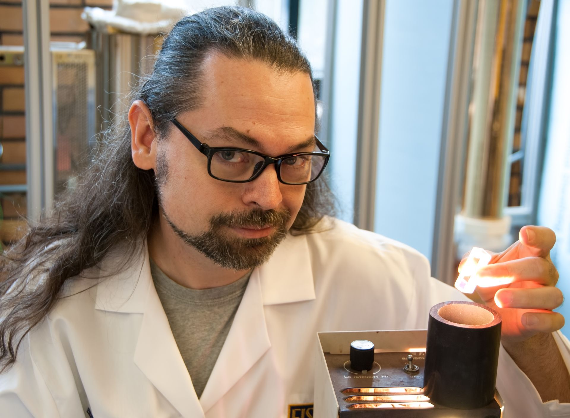 Scientist in a lab coat examines a sample under a microscope, wearing glasses and long hair tied back.