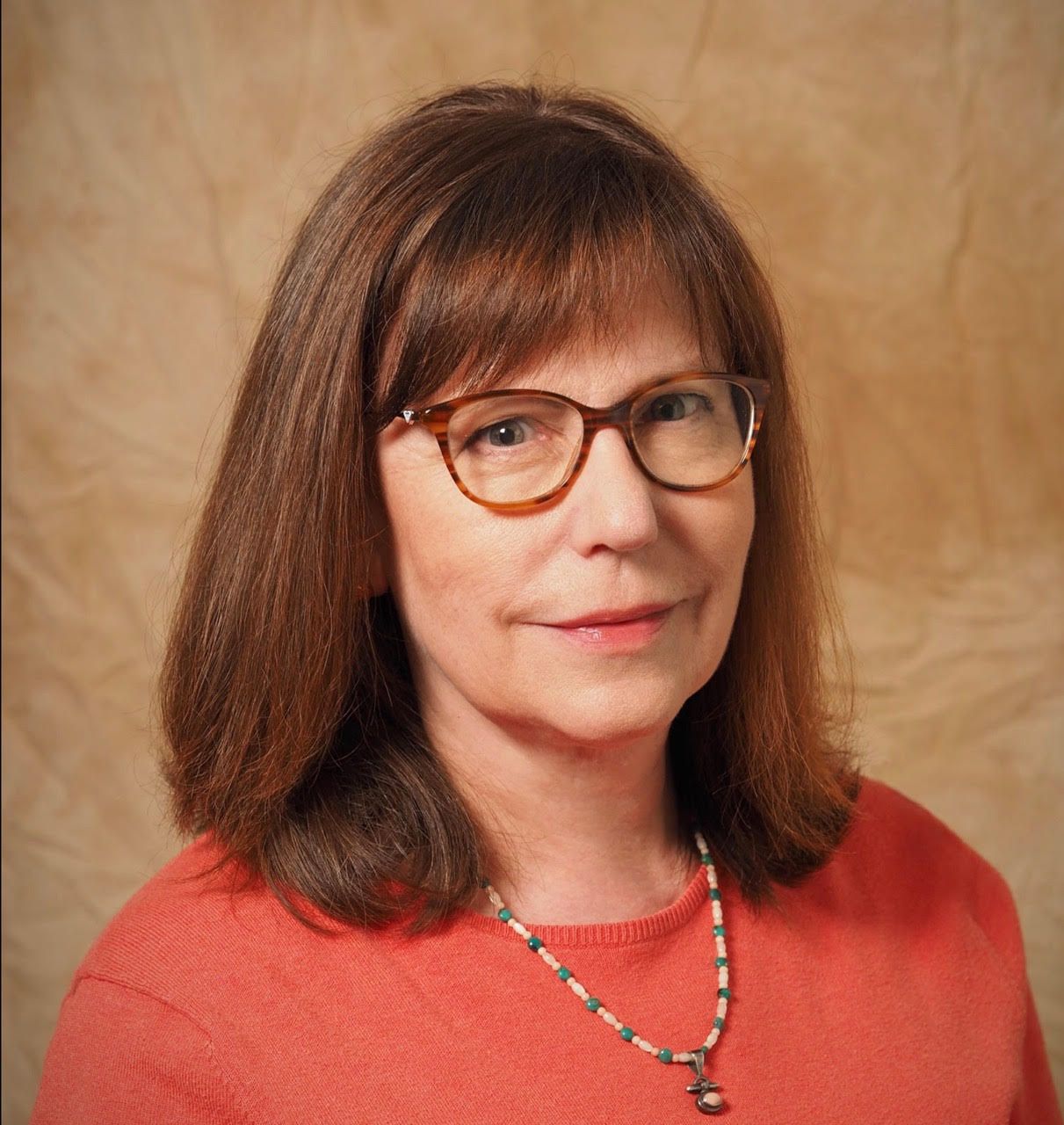 Portrait of a woman with glasses wearing a coral top and necklace against a tan backdrop