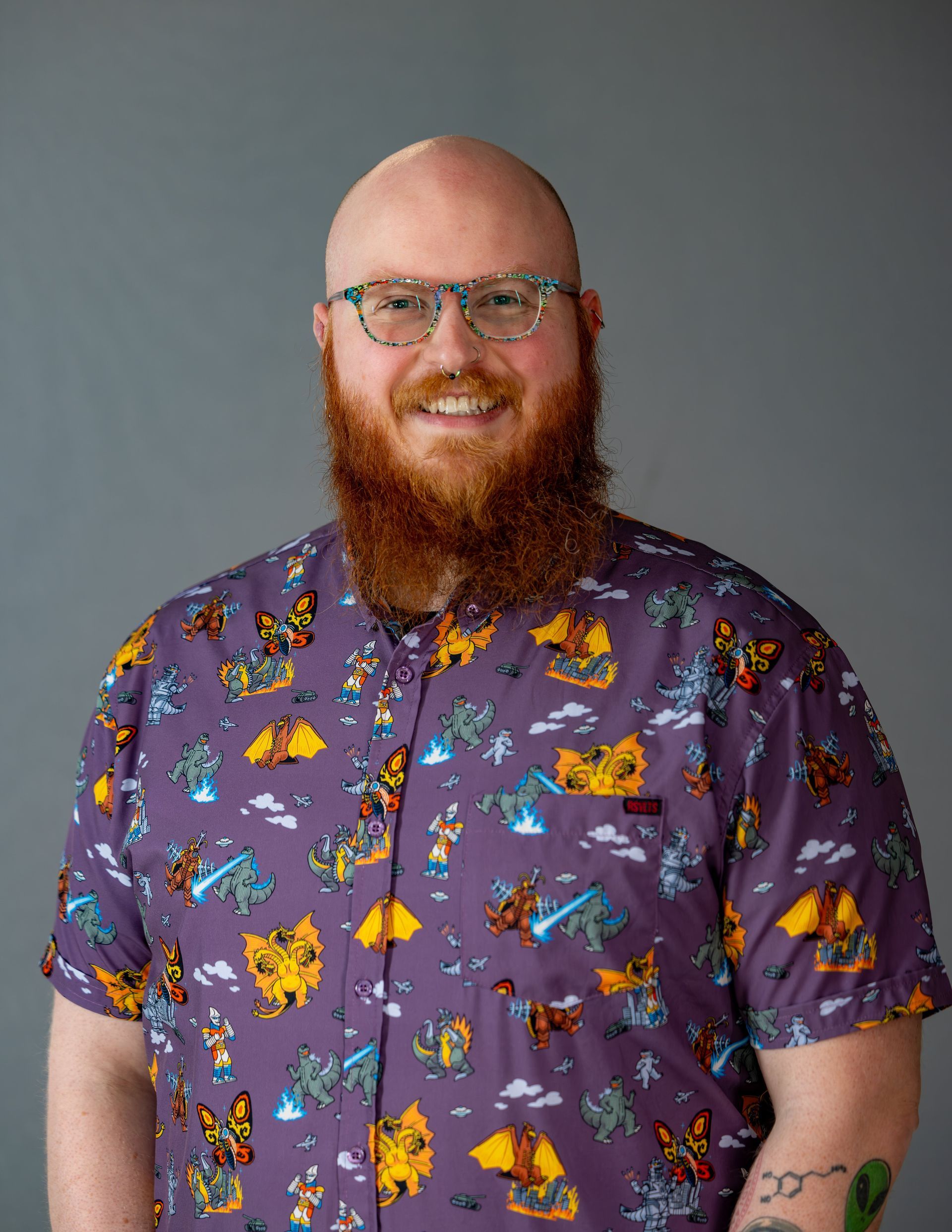 Smiling bald man with a red beard wearing glasses and a purple patterned shirt against a gray background