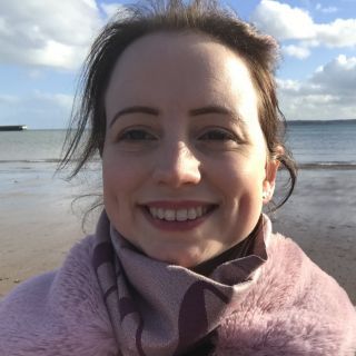 Smiling person wrapped in a pink scarf on a windy beach with cloudy sky and calm sea