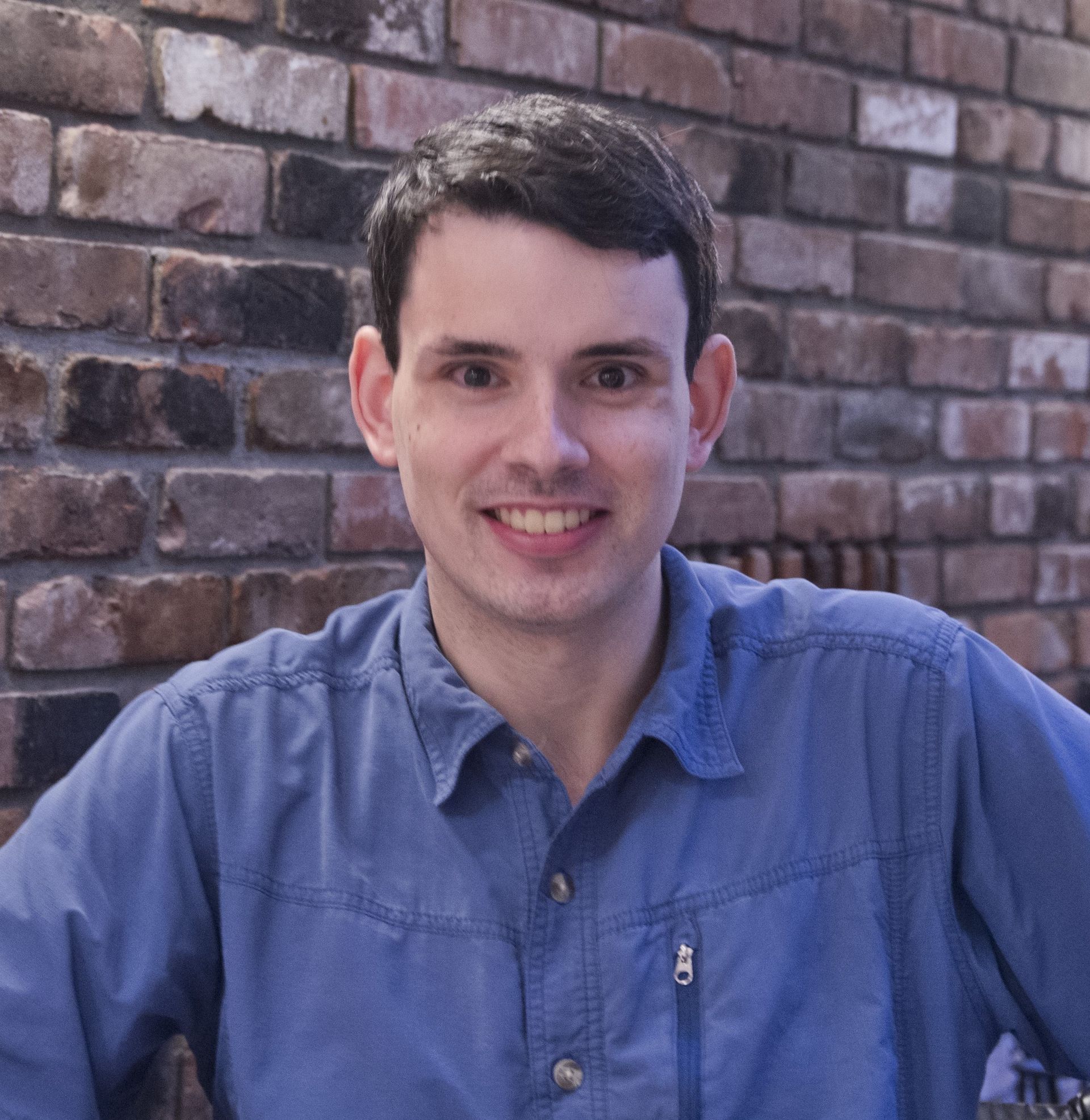 Smiling person in a blue shirt seated in front of a brick wall