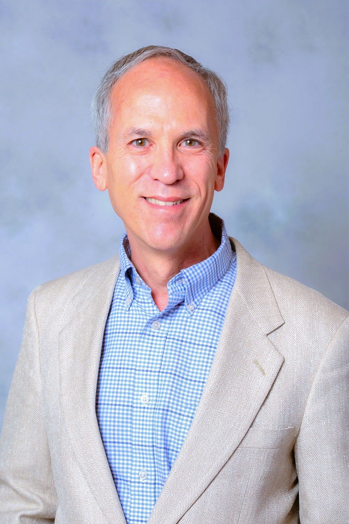 Smiling man in a light gray blazer and blue checkered shirt against a soft blue background