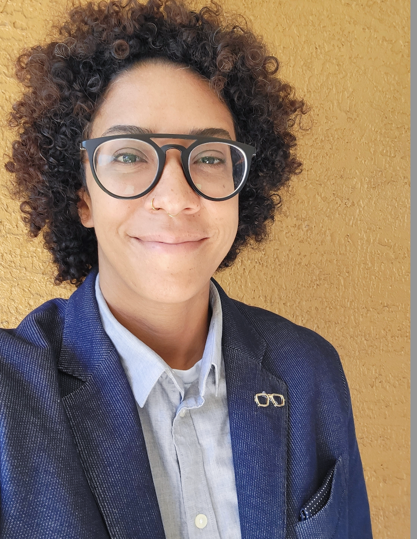 Person smiling in glasses and blue jacket against a yellow wall.