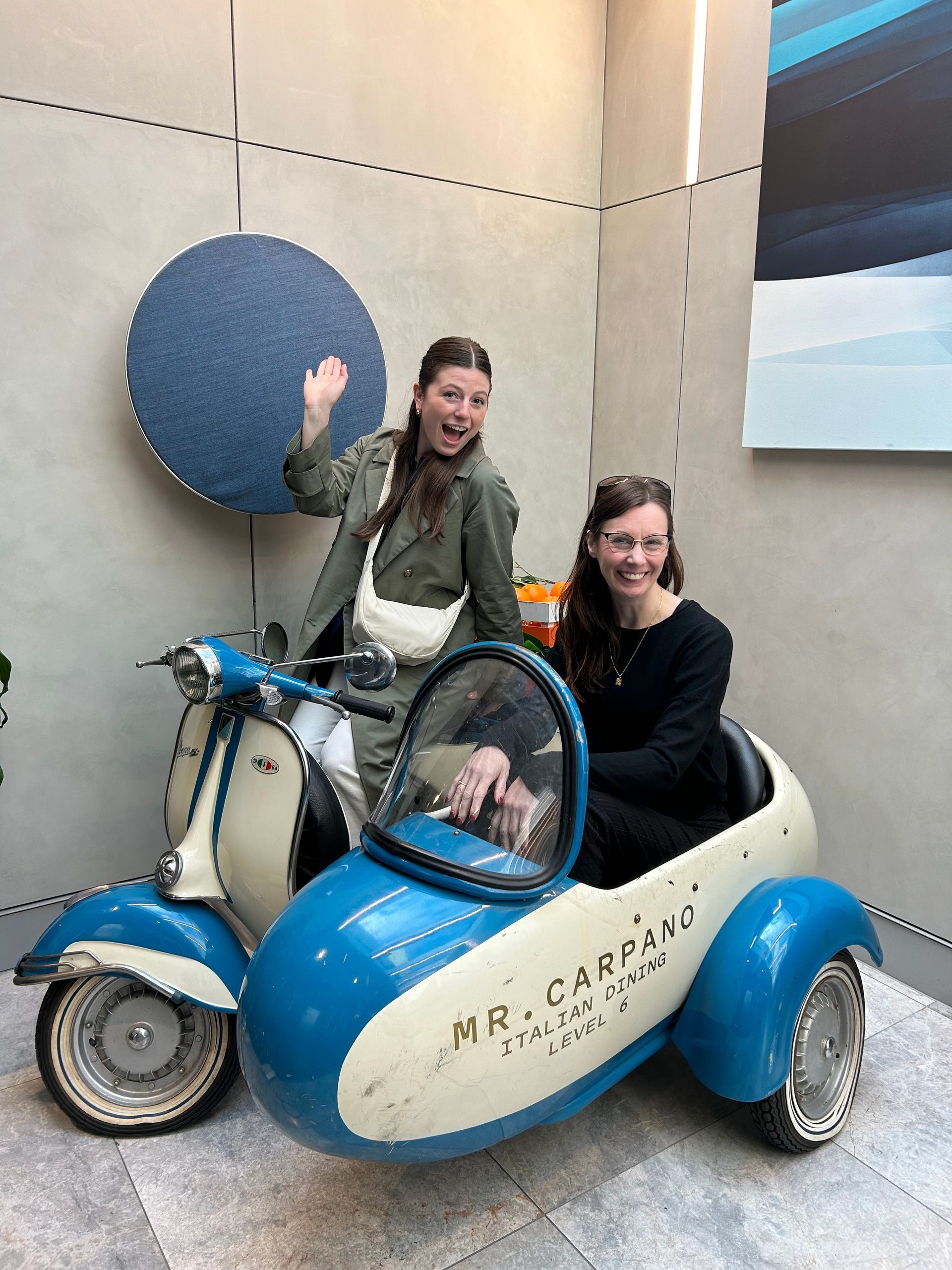 Two people smiling in a blue-and-white vintage toy car indoors, one waving beside a round wall decoration