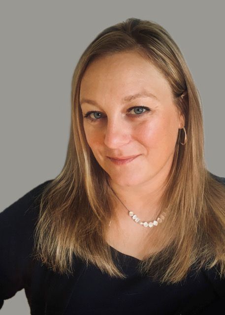Portrait of a woman with long blonde hair wearing a black top and necklace against a gray background