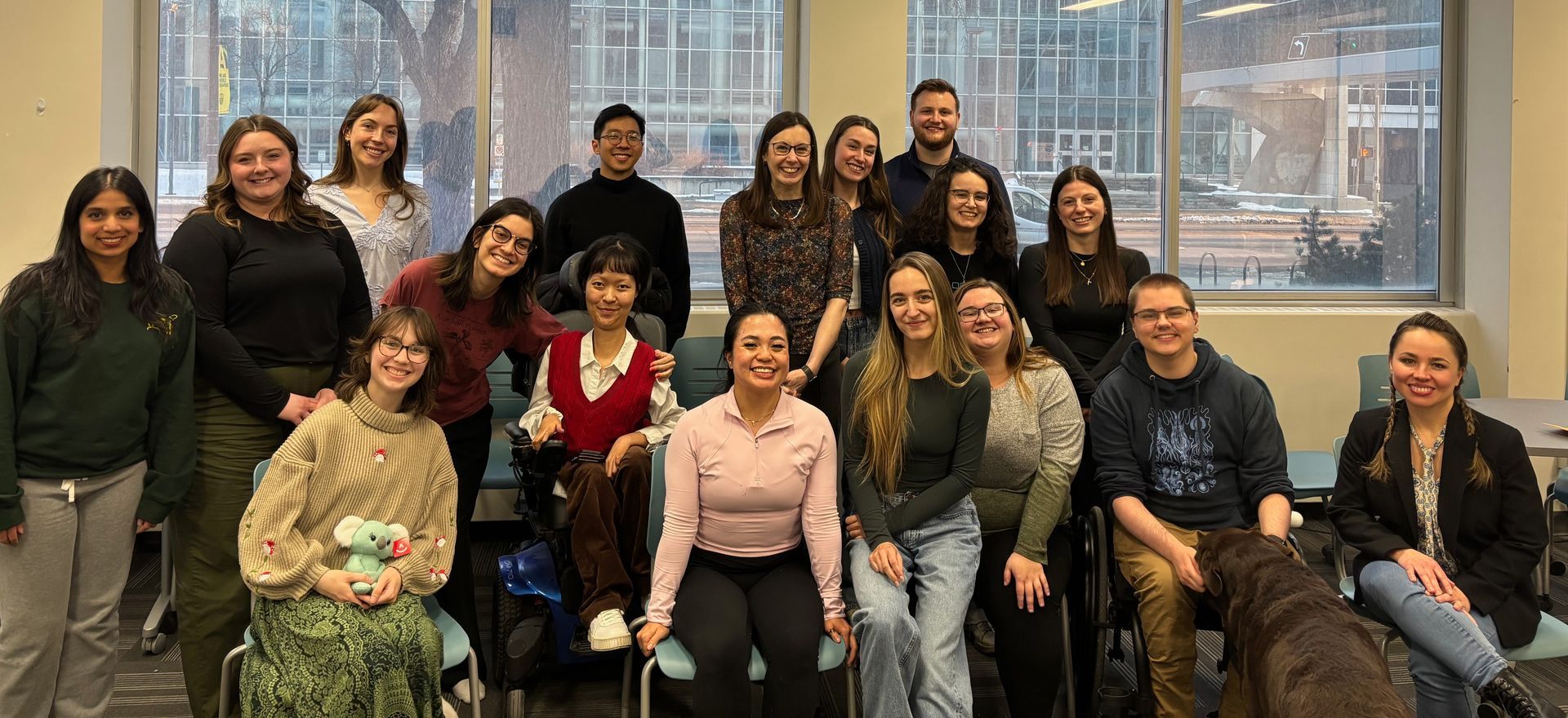 Group of people posing together indoors in front of large windows, smiling in a casual office setting.