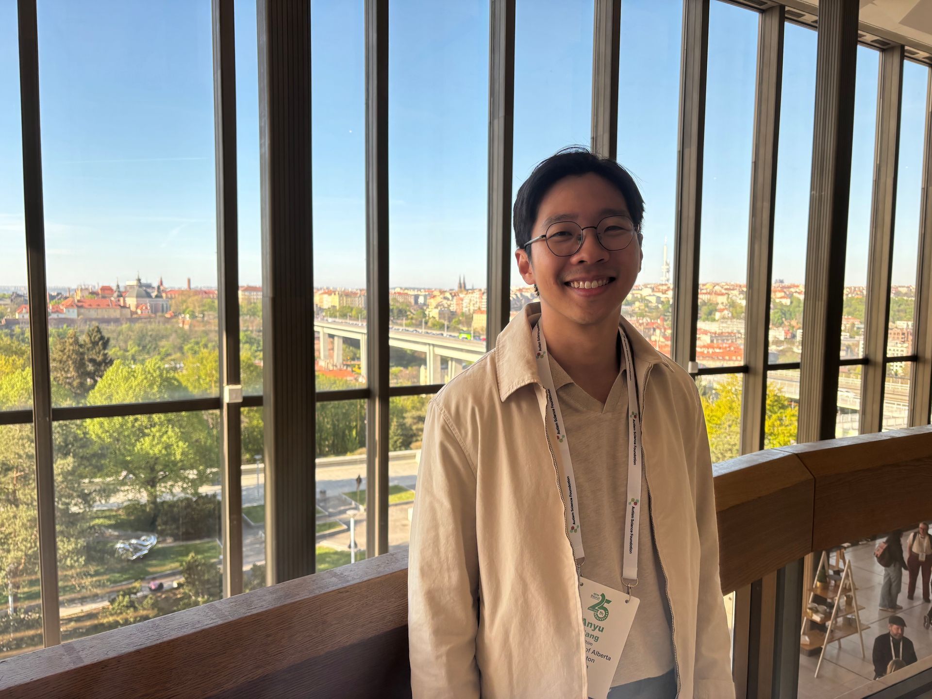 Smiling person in a beige shirt standing by tall window bars with a city view behind them