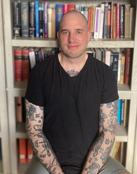 Man seated in front of a bookshelf, wearing a black T-shirt and showing tattooed arms.