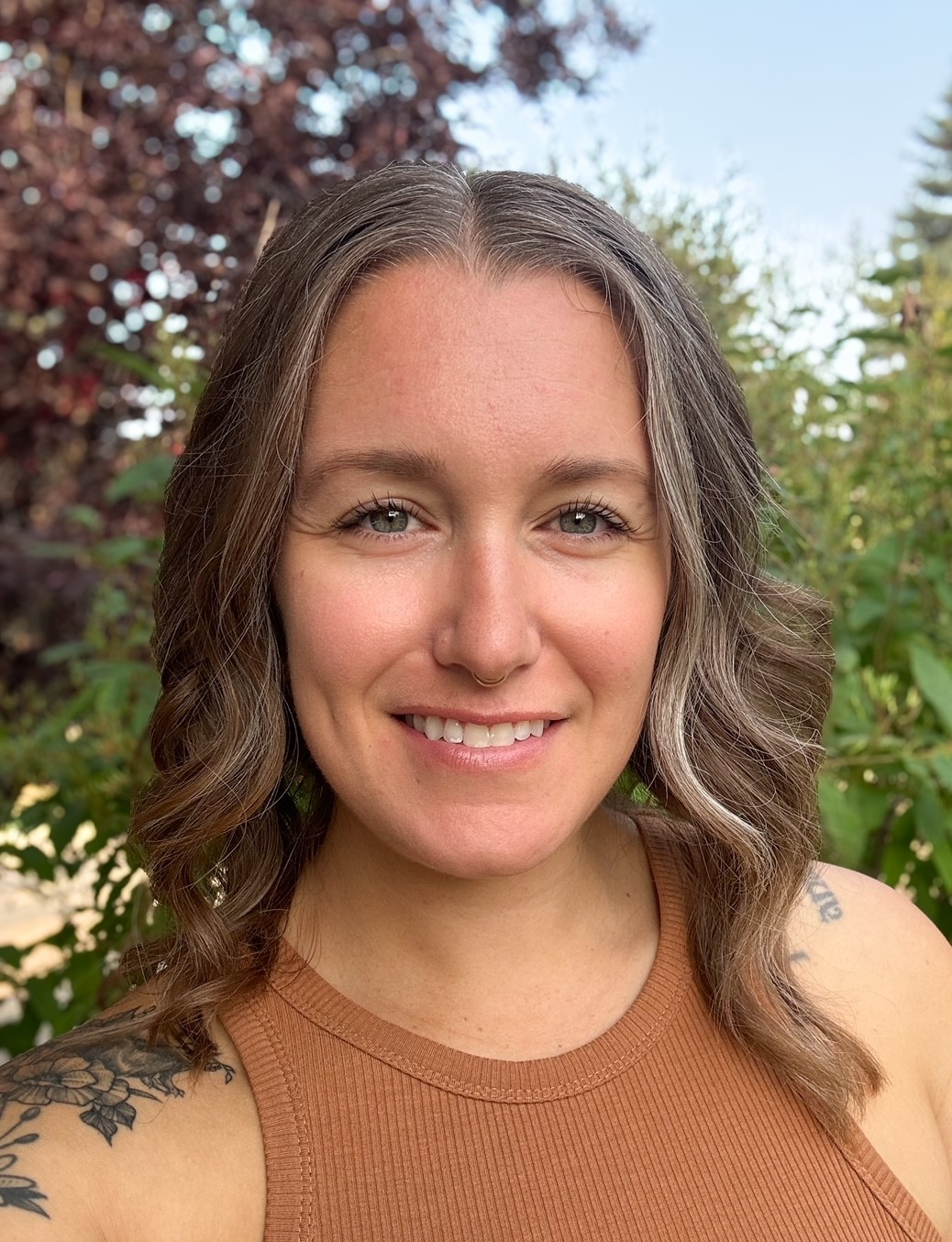 Smiling woman outdoors with curly hair and a brown sleeveless top, standing before greenery.