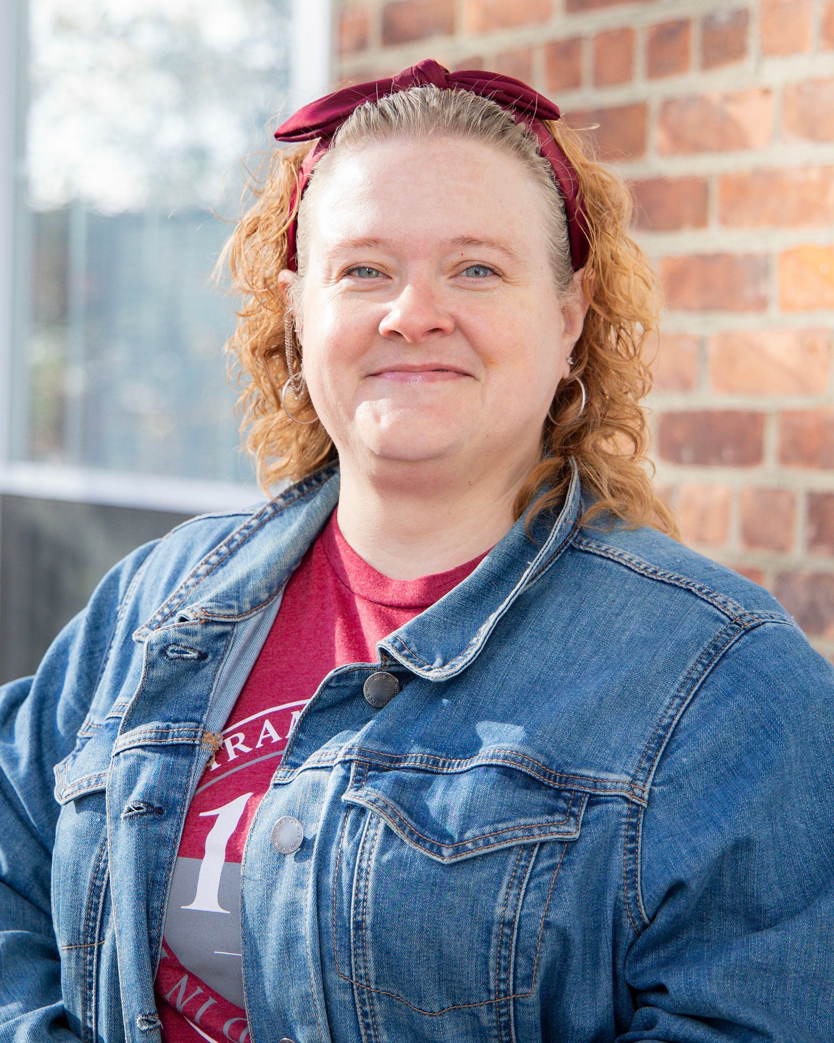 Person in a denim jacket and red shirt standing by a brick wall, smiling at the camera.