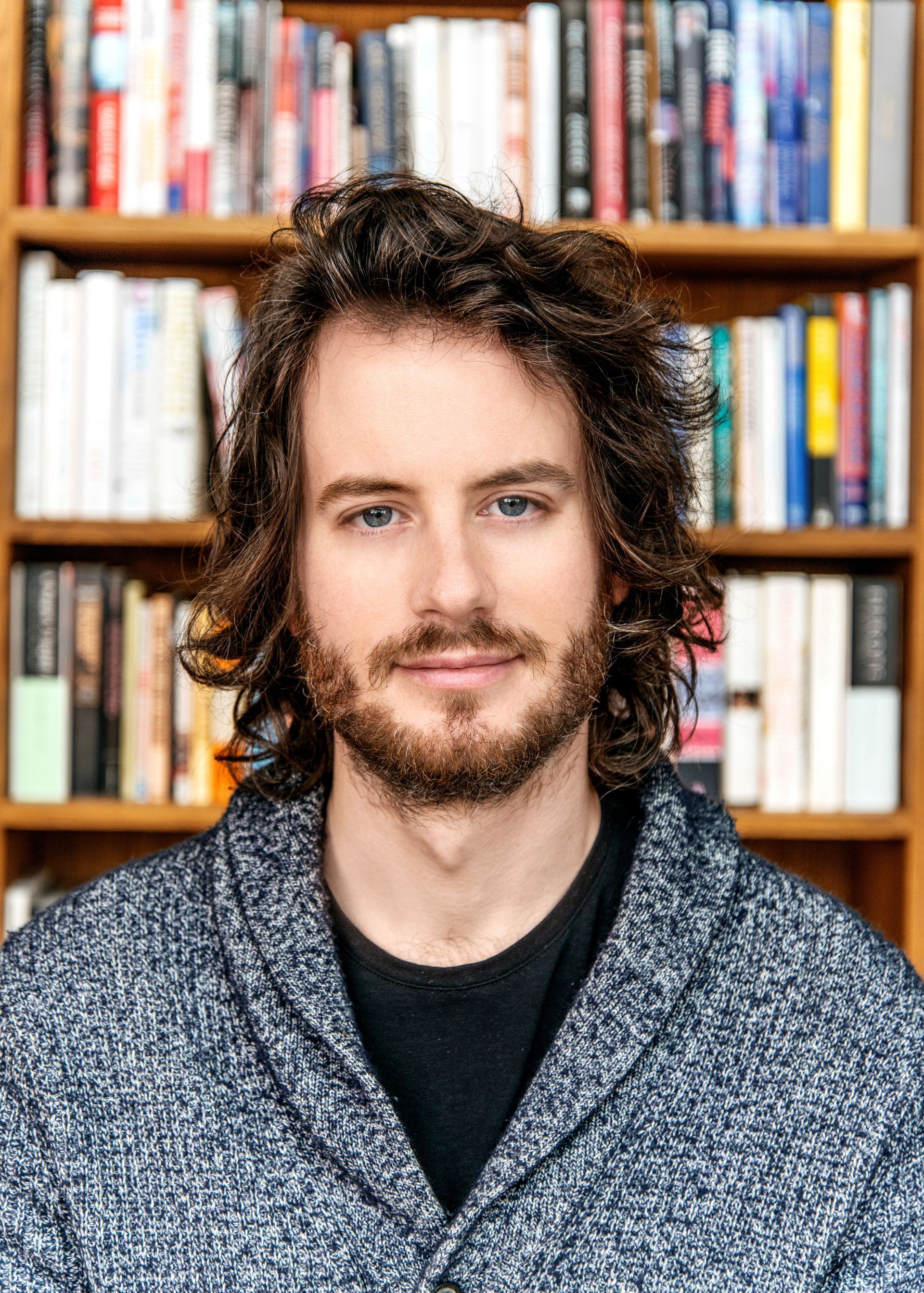 Portrait of a man with curly hair and beard in front of a bookshelf