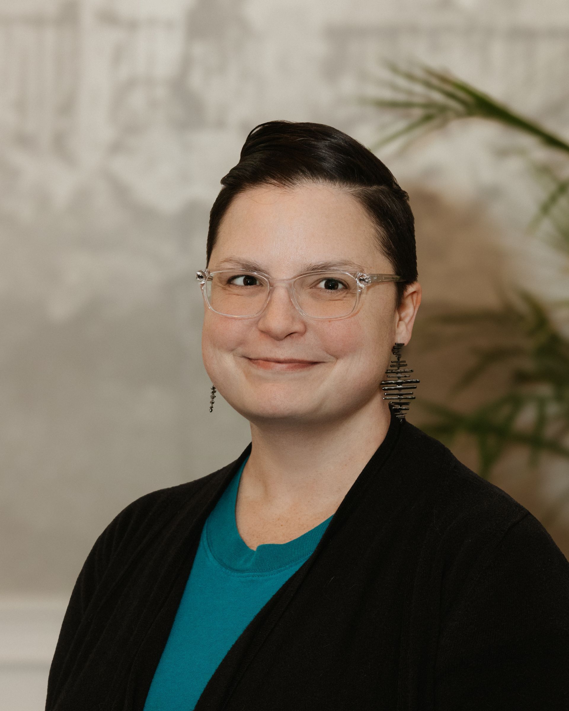 Portrait of a person wearing glasses, a teal shirt, and a black cardigan against a neutral background