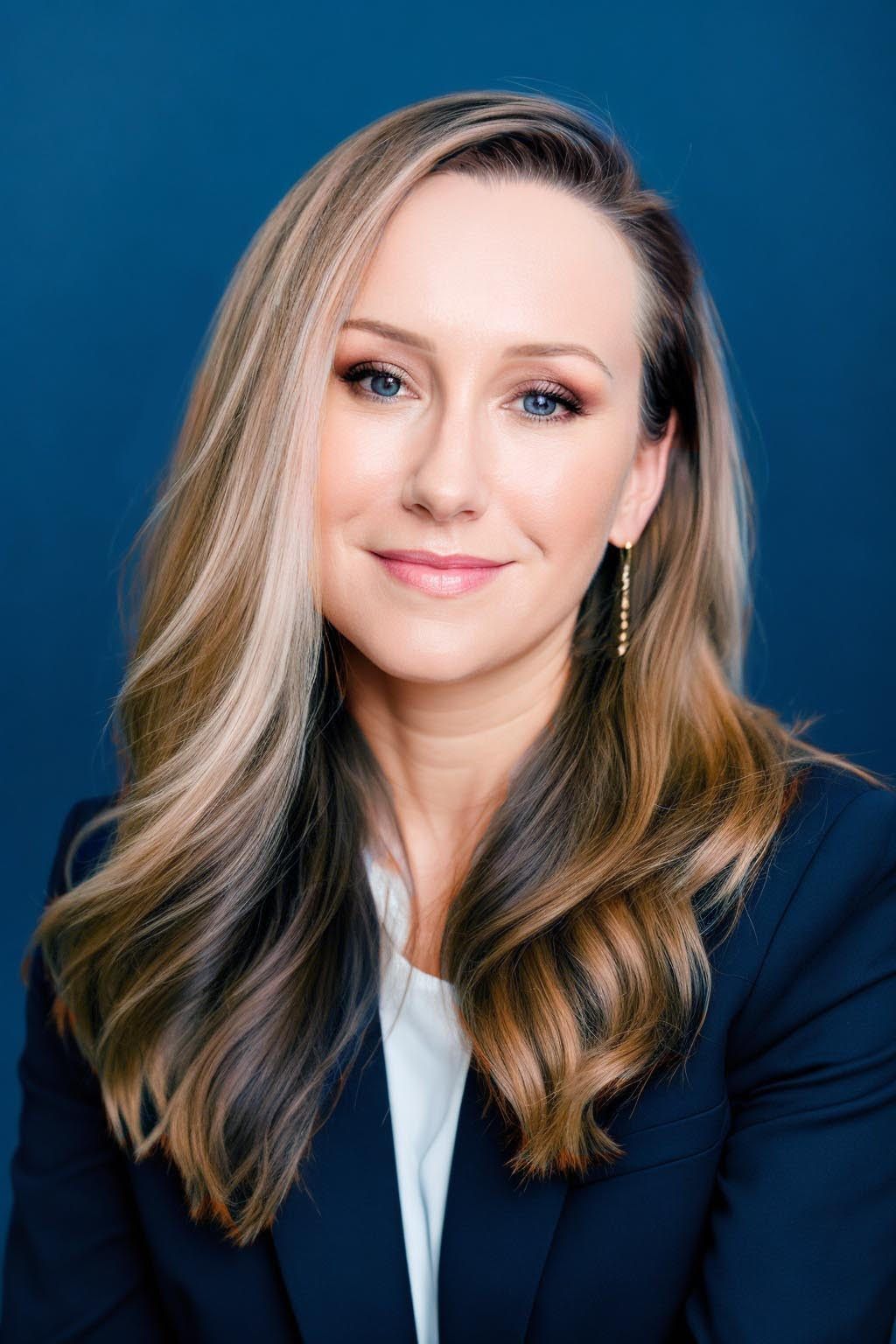 Professional headshot of a smiling person with long blonde hair in a navy blazer against a blue background