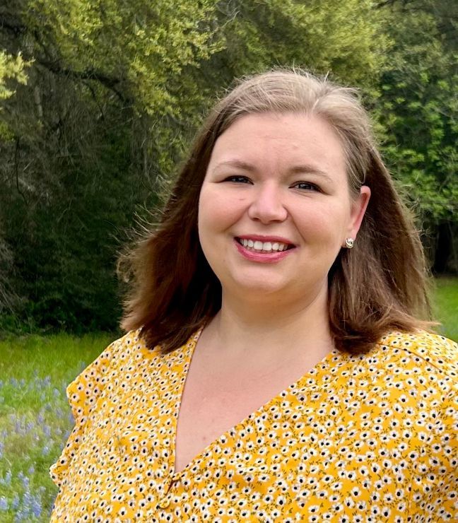 Woman smiling outdoors in a yellow floral top, standing before green trees and grass.