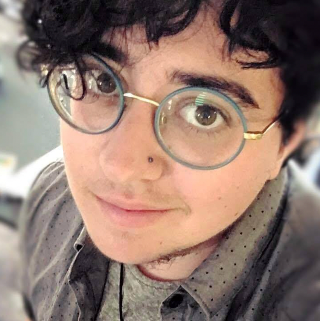Close-up of a person with round glasses and curly dark hair, wearing a gray shirt and smiling softly