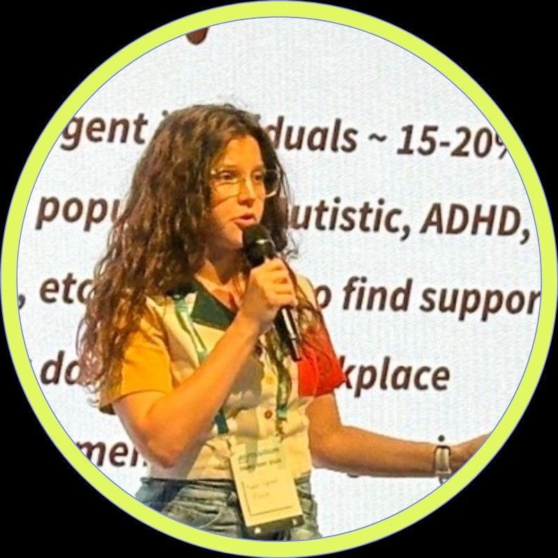 Woman speaking into a microphone on stage, in front of a projected slide about support groups for neurodivergent adults.