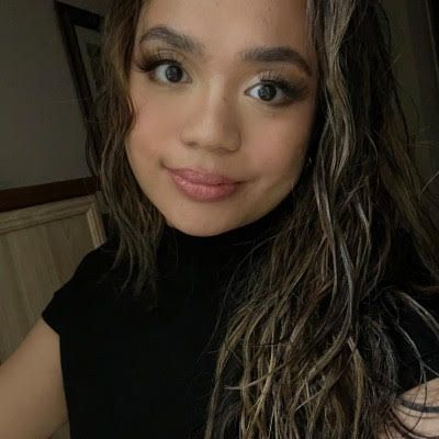 Close-up selfie of a person with wavy highlighted hair and a black top, smiling softly indoors