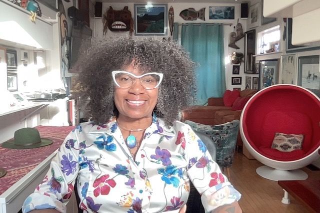 Smiling person in white glasses and floral shirt seated in a bright living room with red chairs.