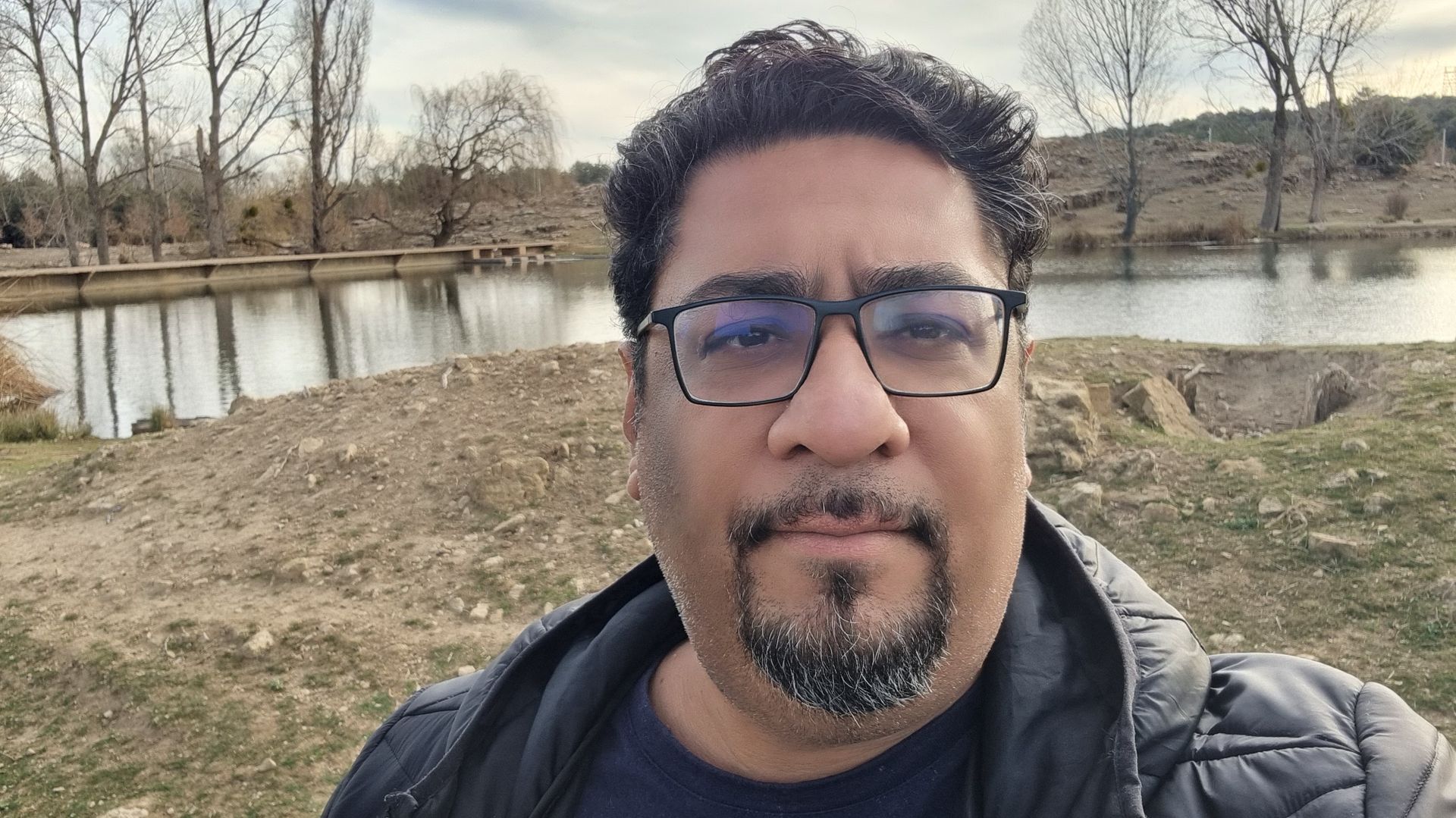 Man taking a selfie by a calm pond with bare trees in the background