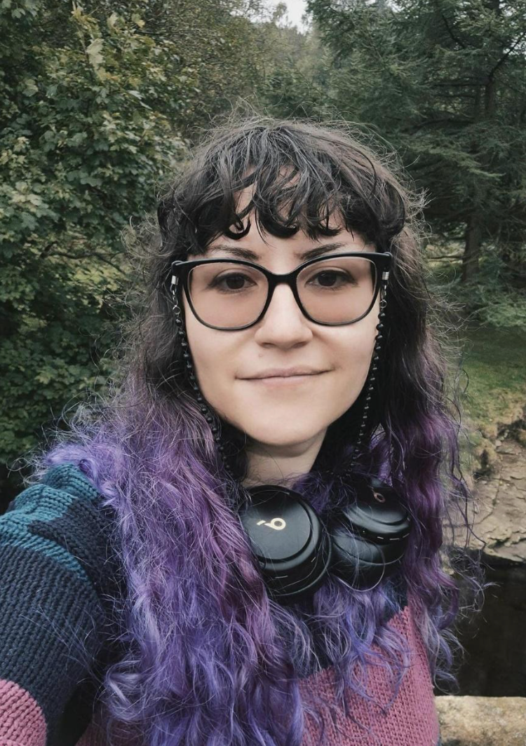 Person with glasses and purple-tinted hair outdoors, wearing headphones, in front of green trees.