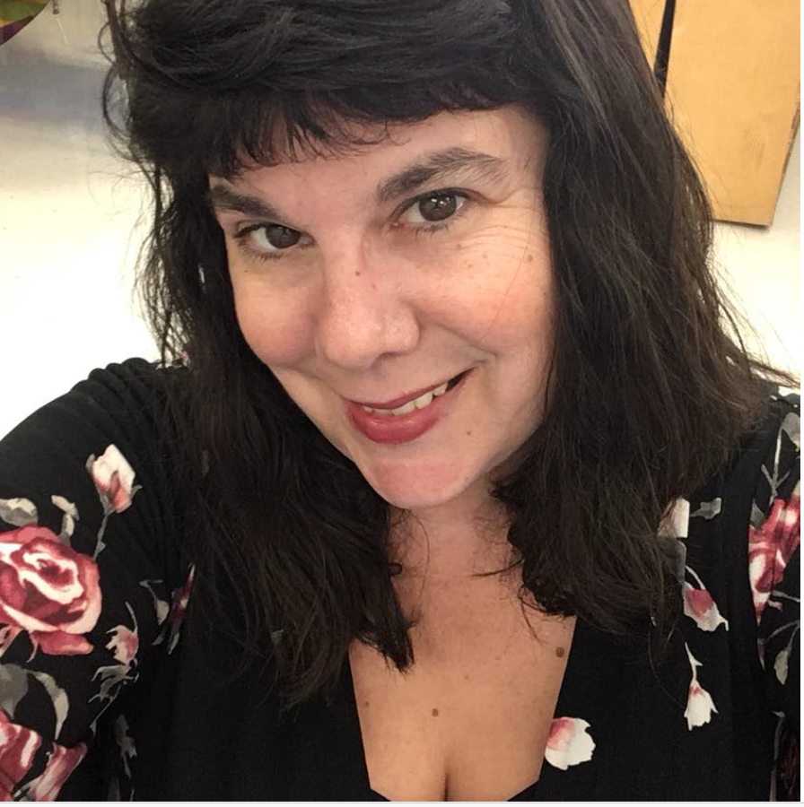 Smiling woman with dark hair wearing a black floral top, taking a selfie indoors