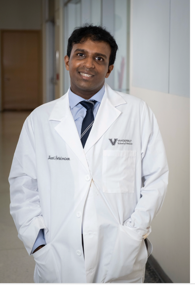 Doctor in a white lab coat standing in a hospital hallway, smiling with hands in pockets.