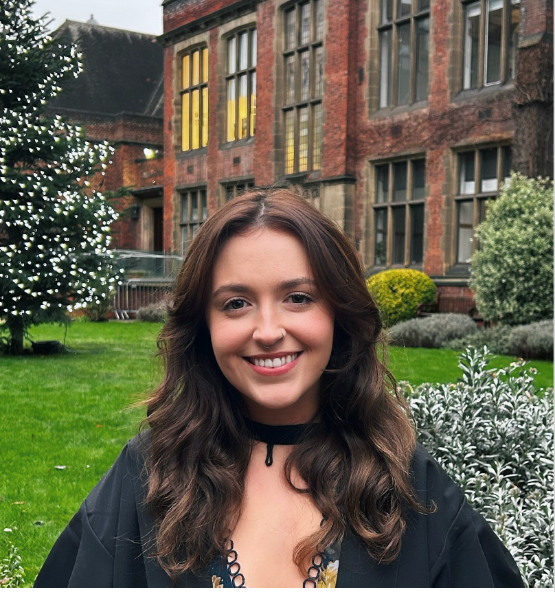 Smiling woman in a black top standing in a garden before a brick building with lit windows