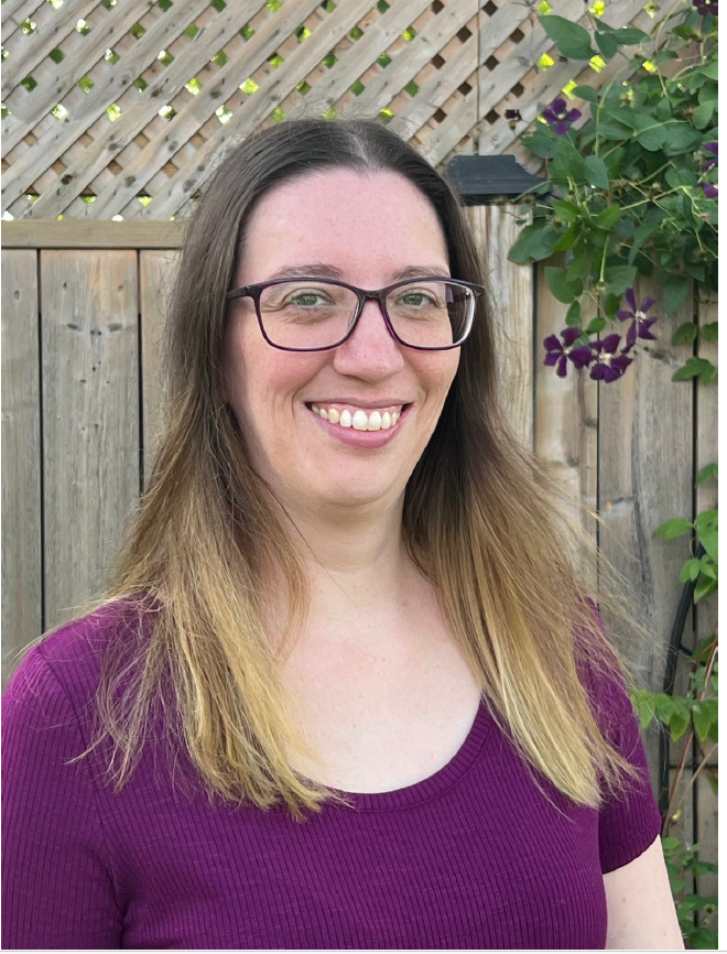 Smiling person with glasses and long hair, wearing a purple top, standing in front of a wooden fence and trellis.