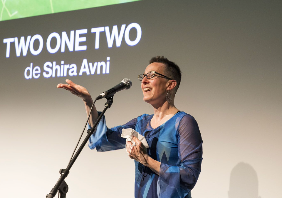 Speaker in blue dress on stage, gesturing at a microphone beside “TWO ONE TWO de Shira Avni” backdrop