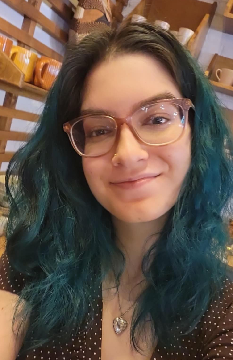 Woman with teal hair and glasses smiling indoors in front of wooden shelves