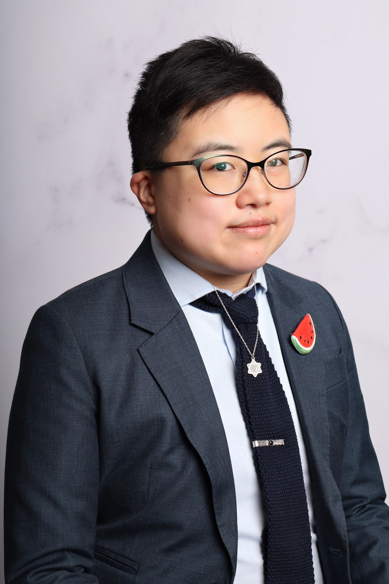 Studio portrait of a person in a dark suit and glasses, wearing a red pin, against a light gray background