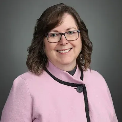 Smiling woman in glasses and a pink blazer against a gray background