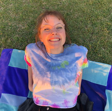 A woman in a tie dye shirt is laying on a blanket in the grass.