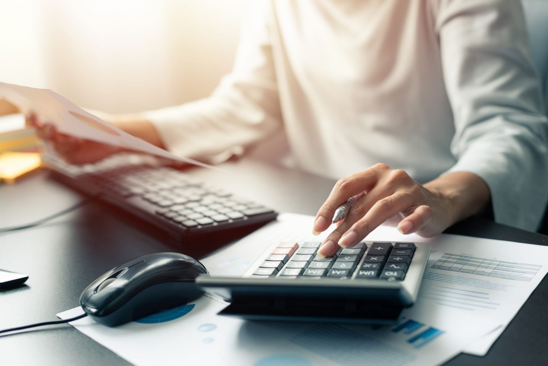 A woman is typing on a calculator while holding a piece of paper.