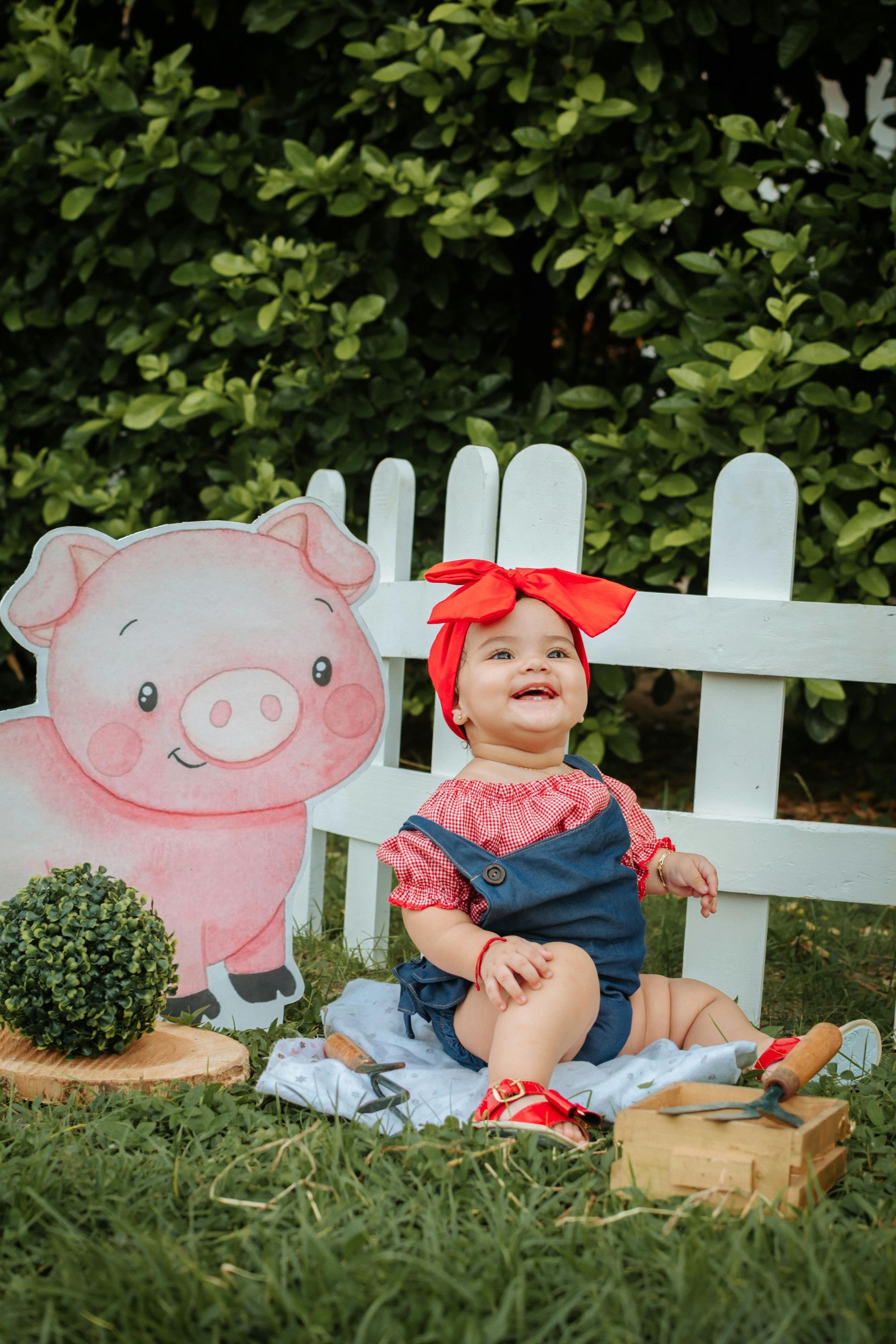 Niña sentada junto a una figura de cartón de un cerdo y una valla blanca en un jardín con césped