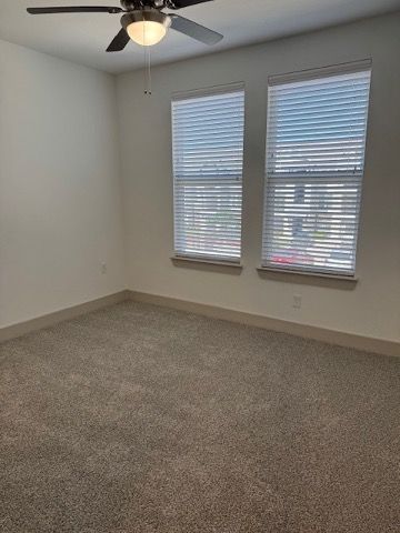 Empty carpeted bedroom with white walls, two windows with blinds, and a ceiling fan.