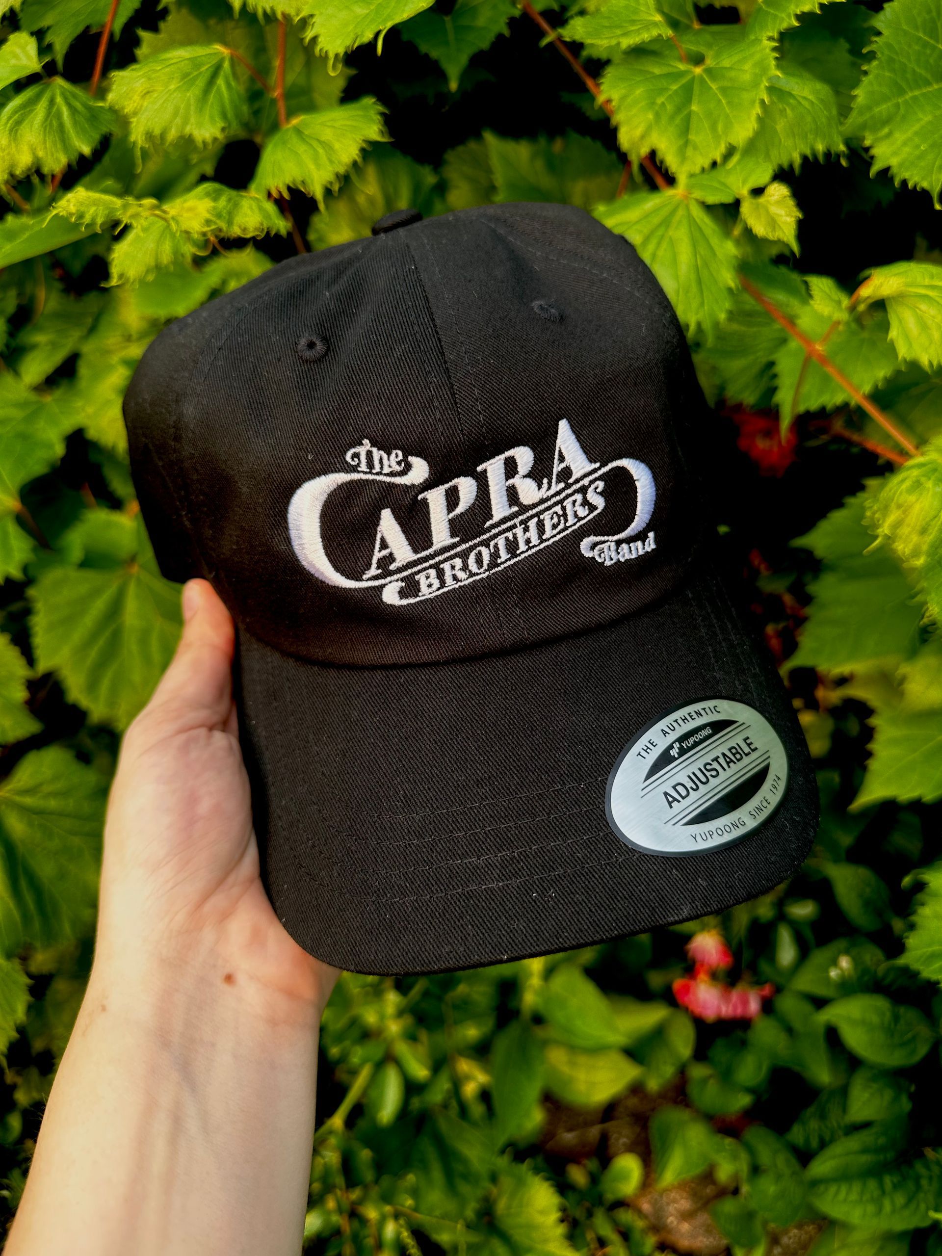 A person is holding a black cap in front of a bush.