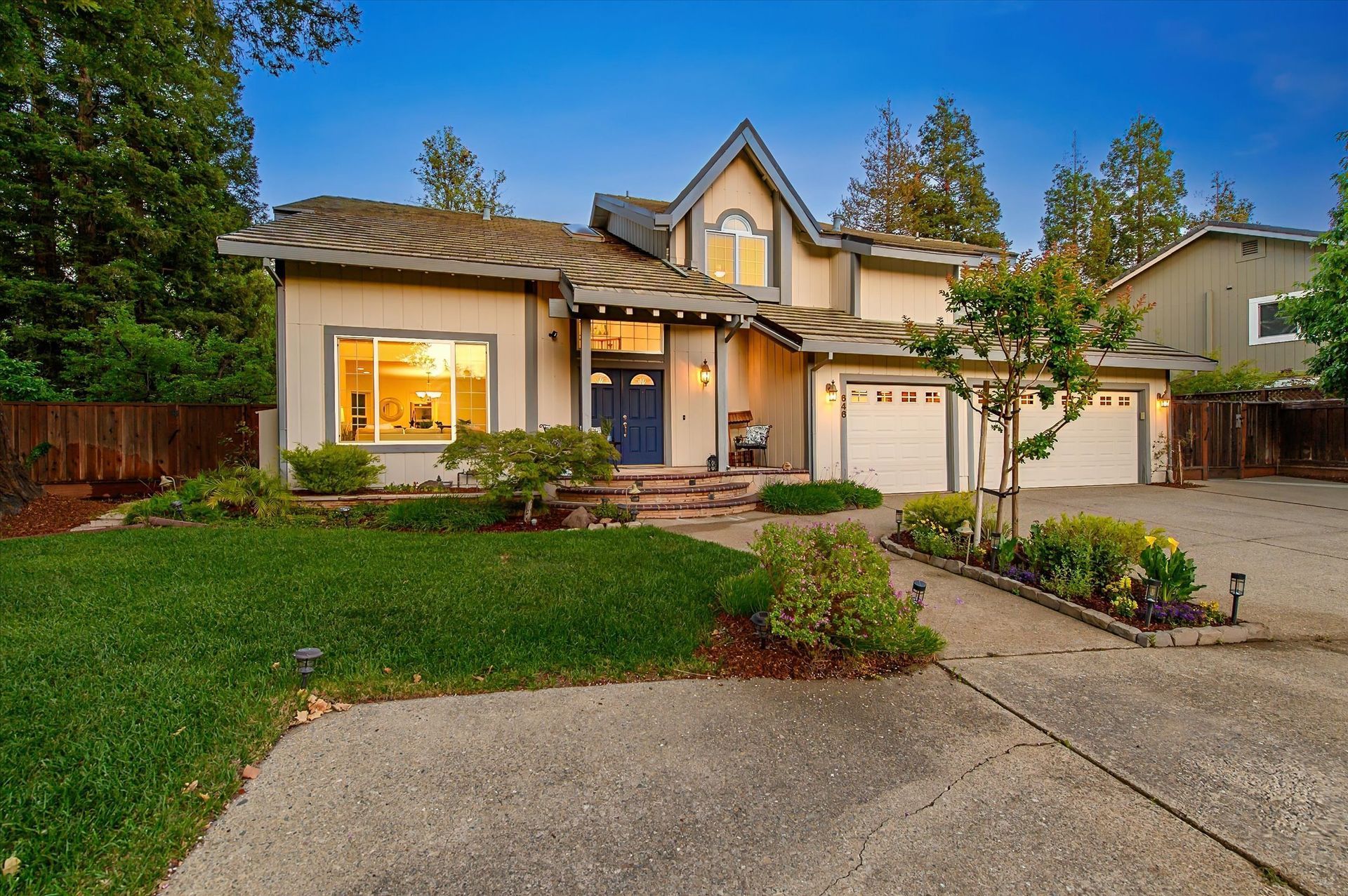 Suburban two-story house with lit windows, front lawn, driveway, and sunset sky