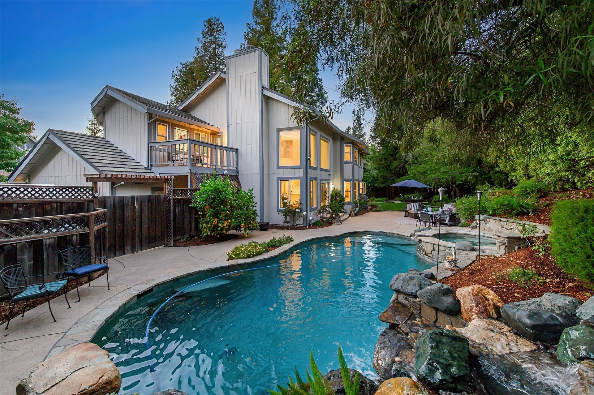 Backyard pool beside a modern house at dusk, with warm lit windows and lush landscaping