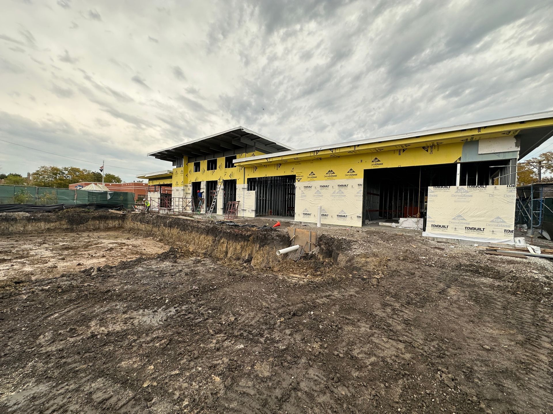 Construction site with building frame, exposed insulation, and muddy ground under cloudy sky.