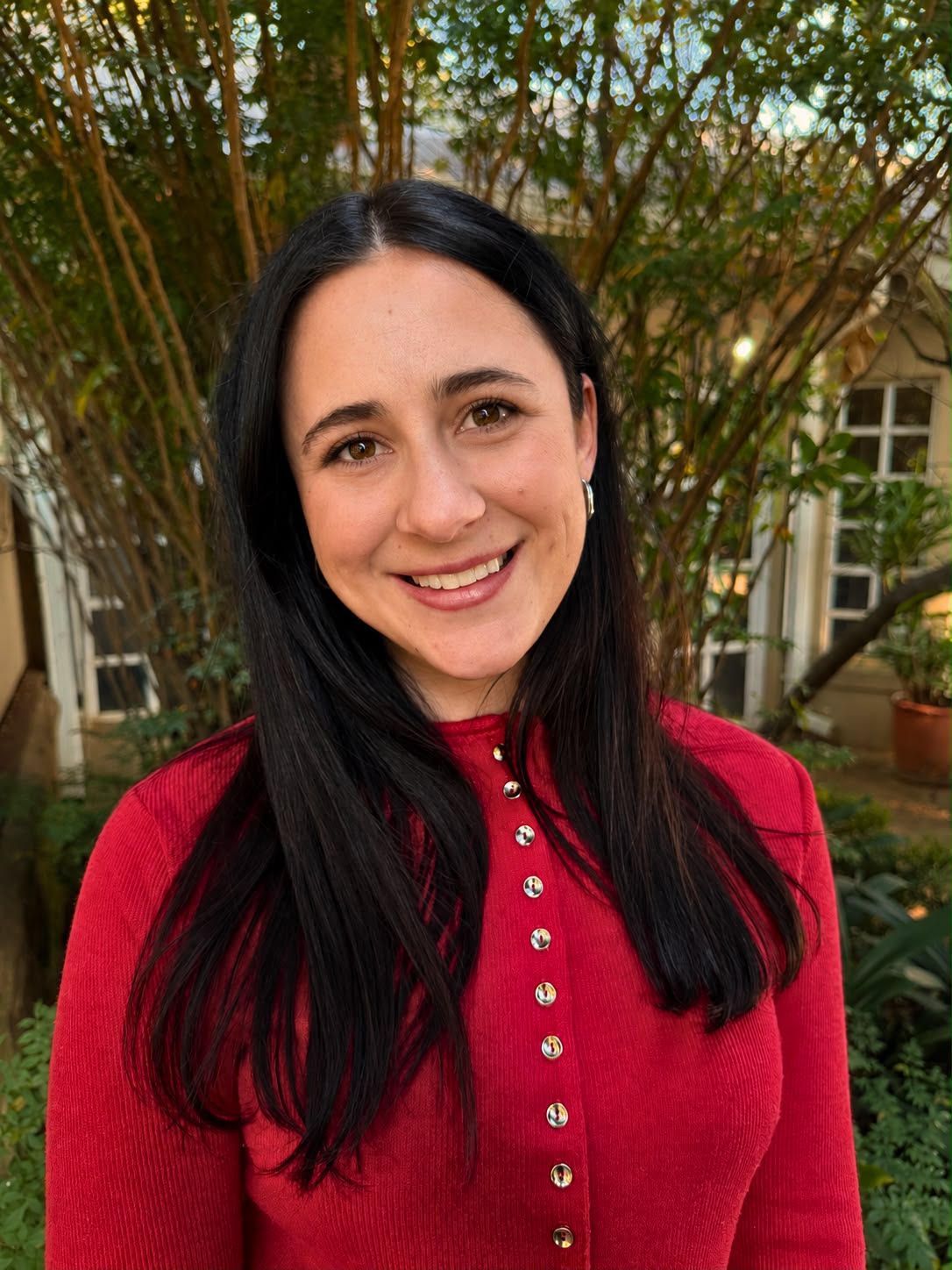 Smiling person in a red sweater standing outdoors in front of trees and a building.