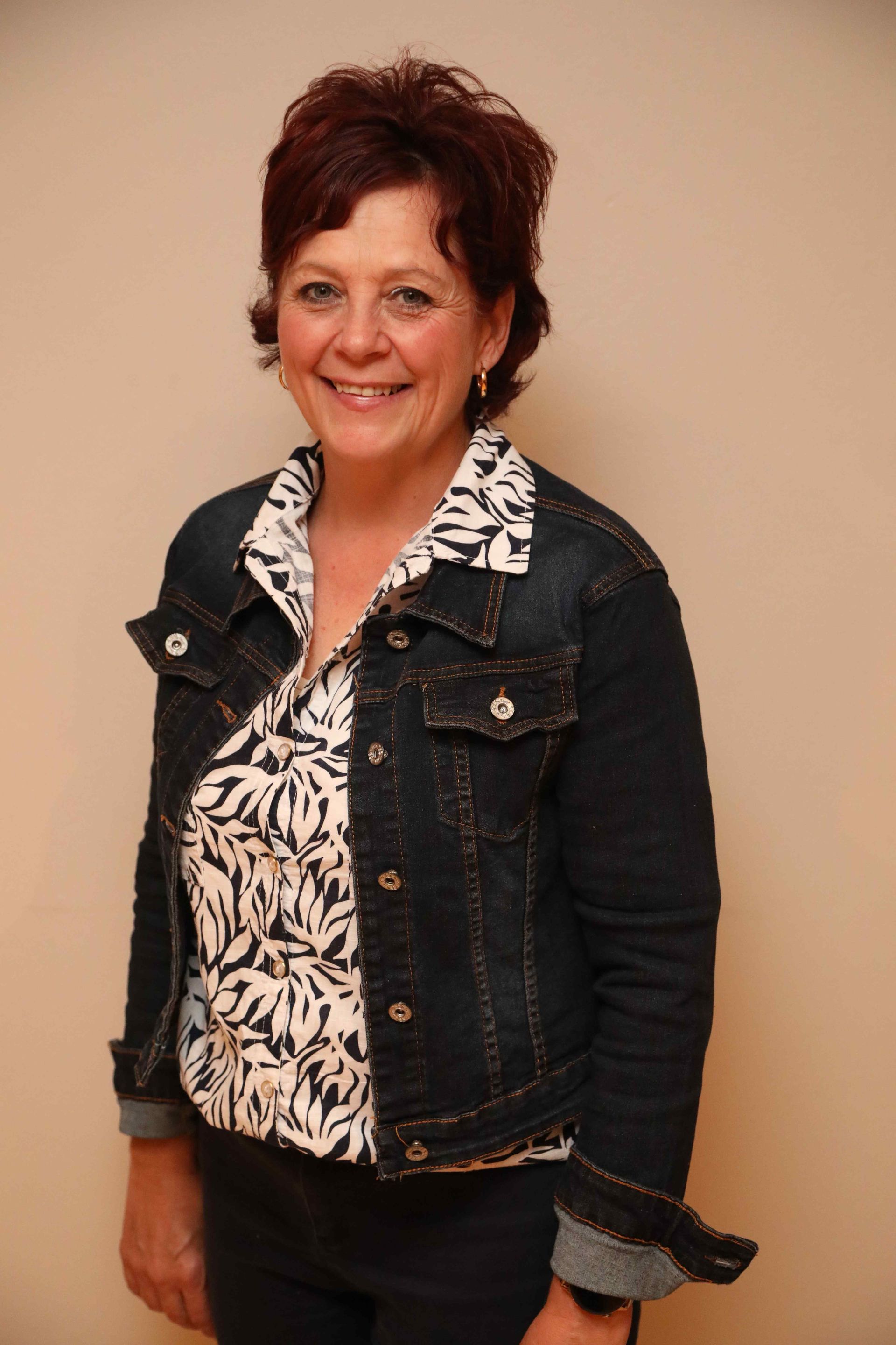 Smiling person in a dark denim jacket over a patterned blouse against a plain beige background