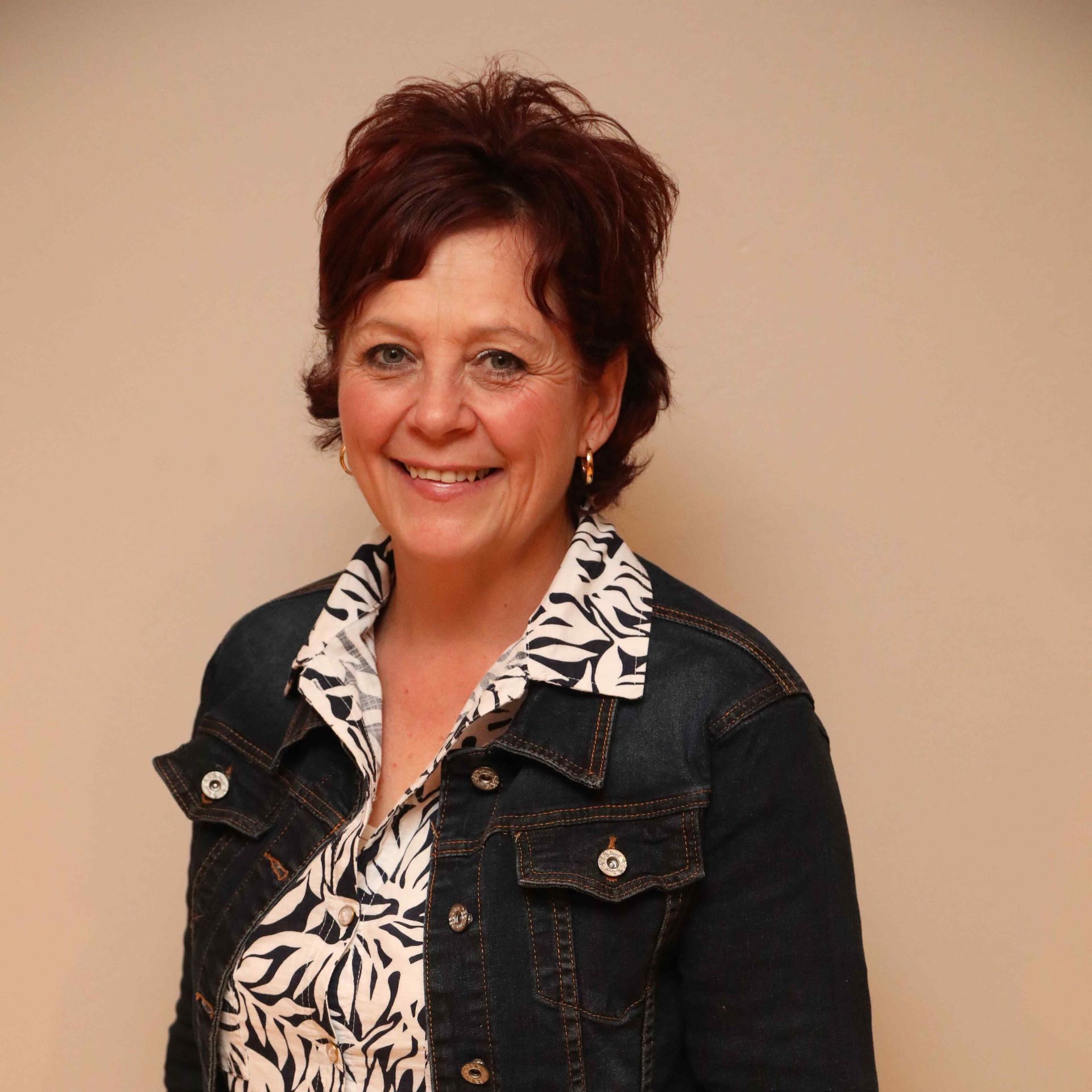 Woman smiling in a denim jacket over a black-and-white patterned blouse against a plain beige background
