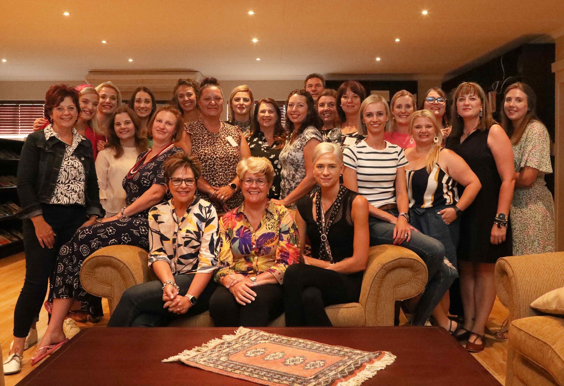 Large group posing together in a warmly lit lounge, seated and standing around sofas and a coffee table.