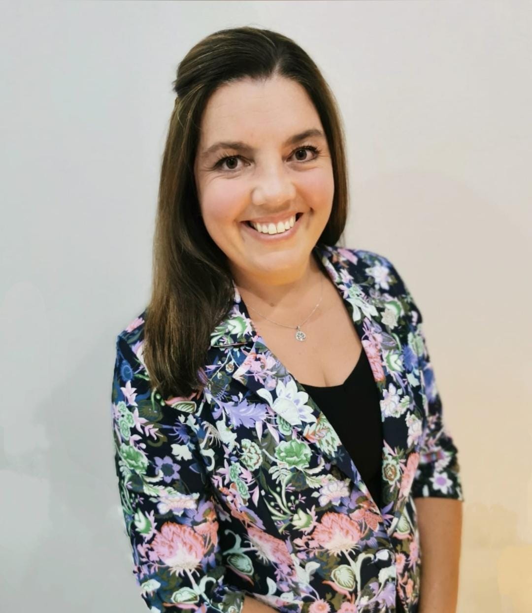 Smiling woman in a floral blazer standing against a light gray background.