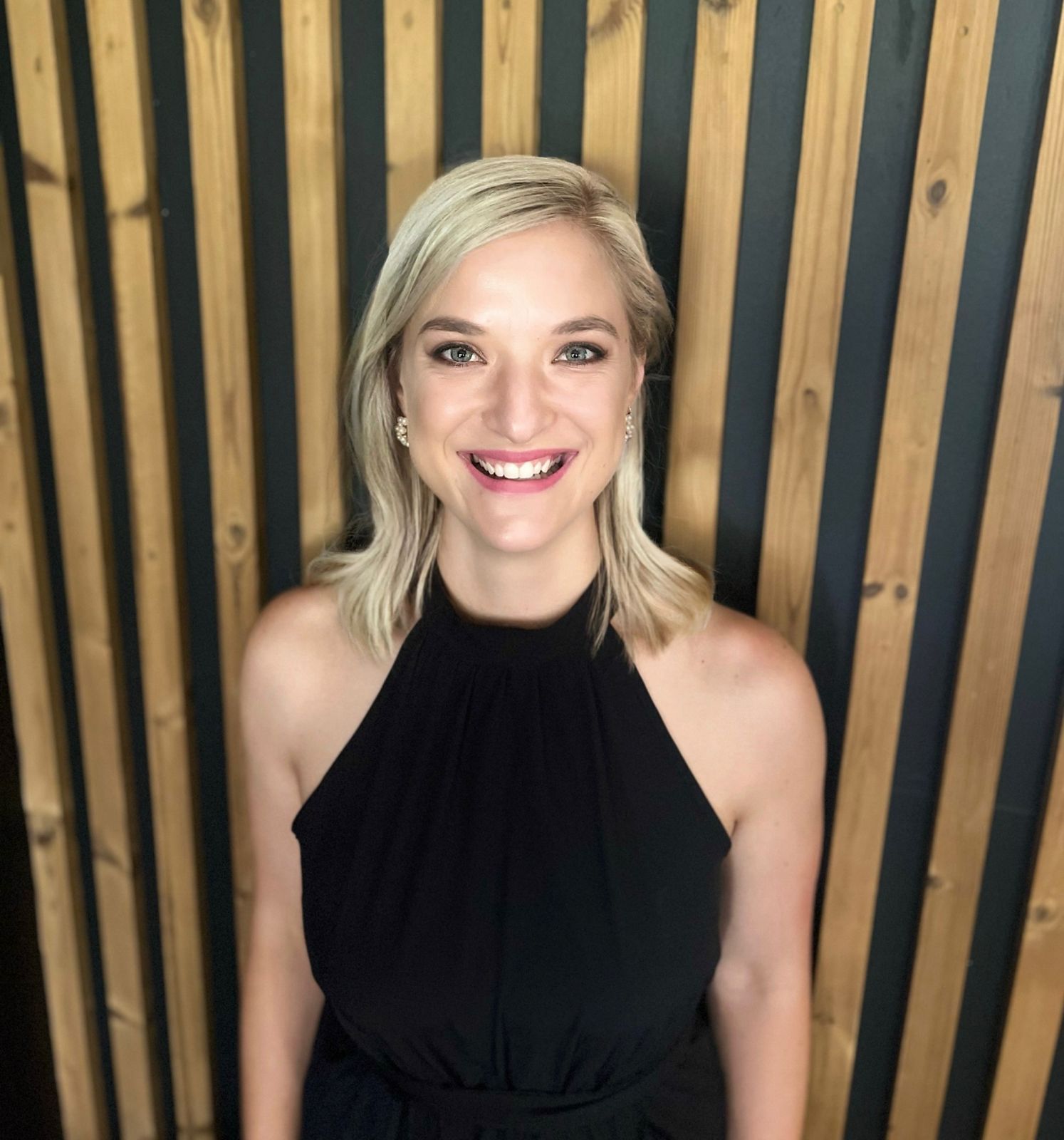 Smiling blonde woman in a black halter top standing before a wood slat wall.